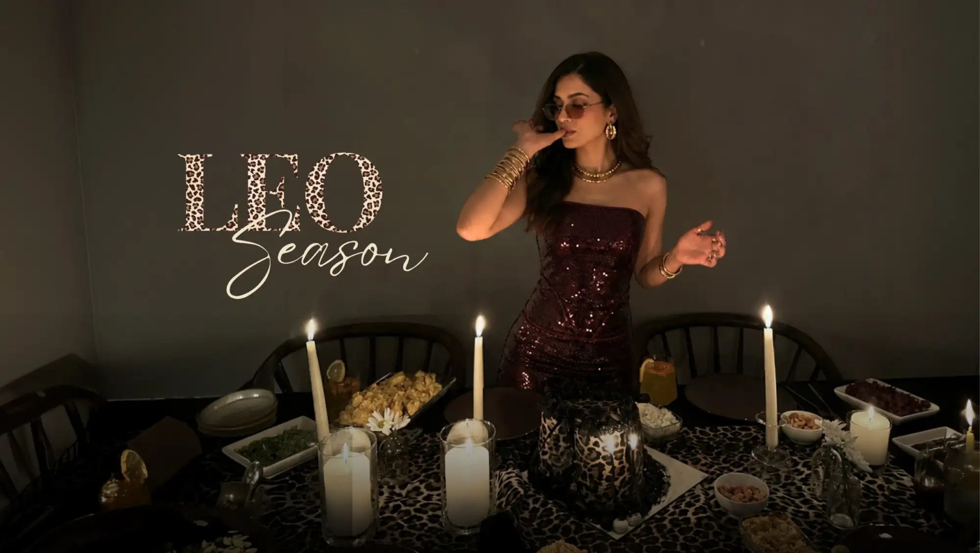A woman in a shimmering, deep burgundy sequined strapless dress stands elegantly beside an ornately set dining table adorned with candles and leopard-print accents.