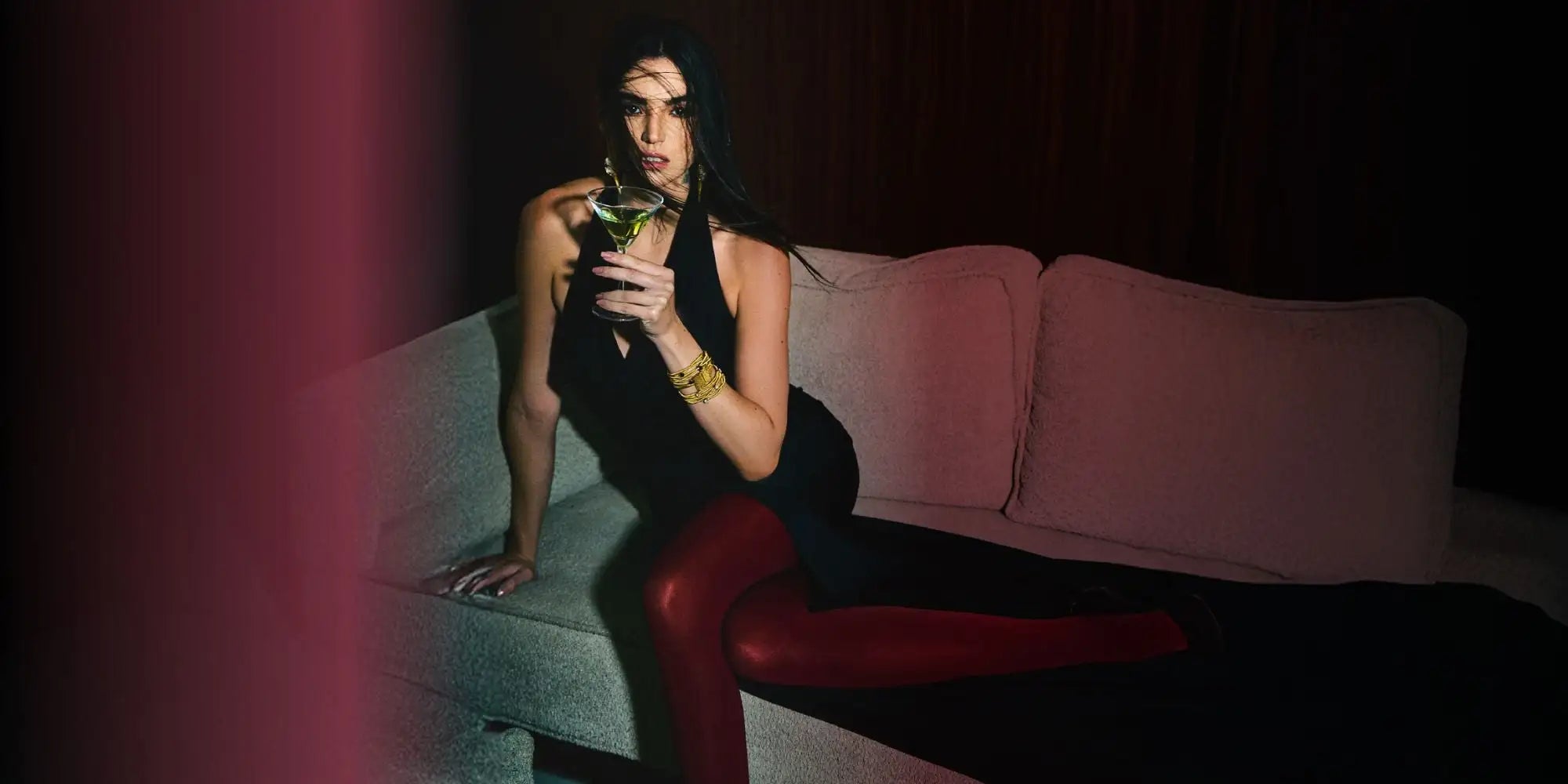 A woman in a sleek black dress and glossy red leggings sits elegantly on a muted gray couch, holding a martini glass.